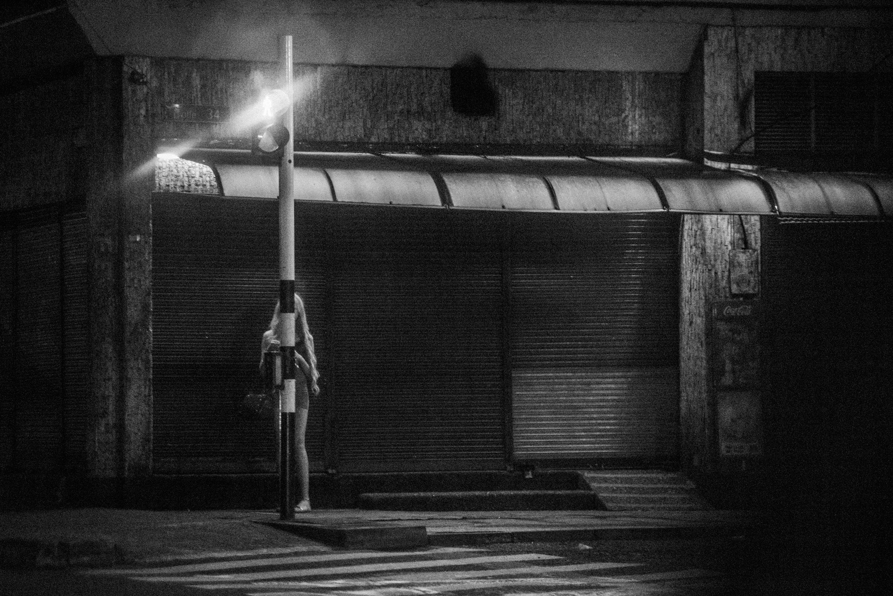 Una figura junto a una farola, frente a persianas cerradas, en blanco y negro.