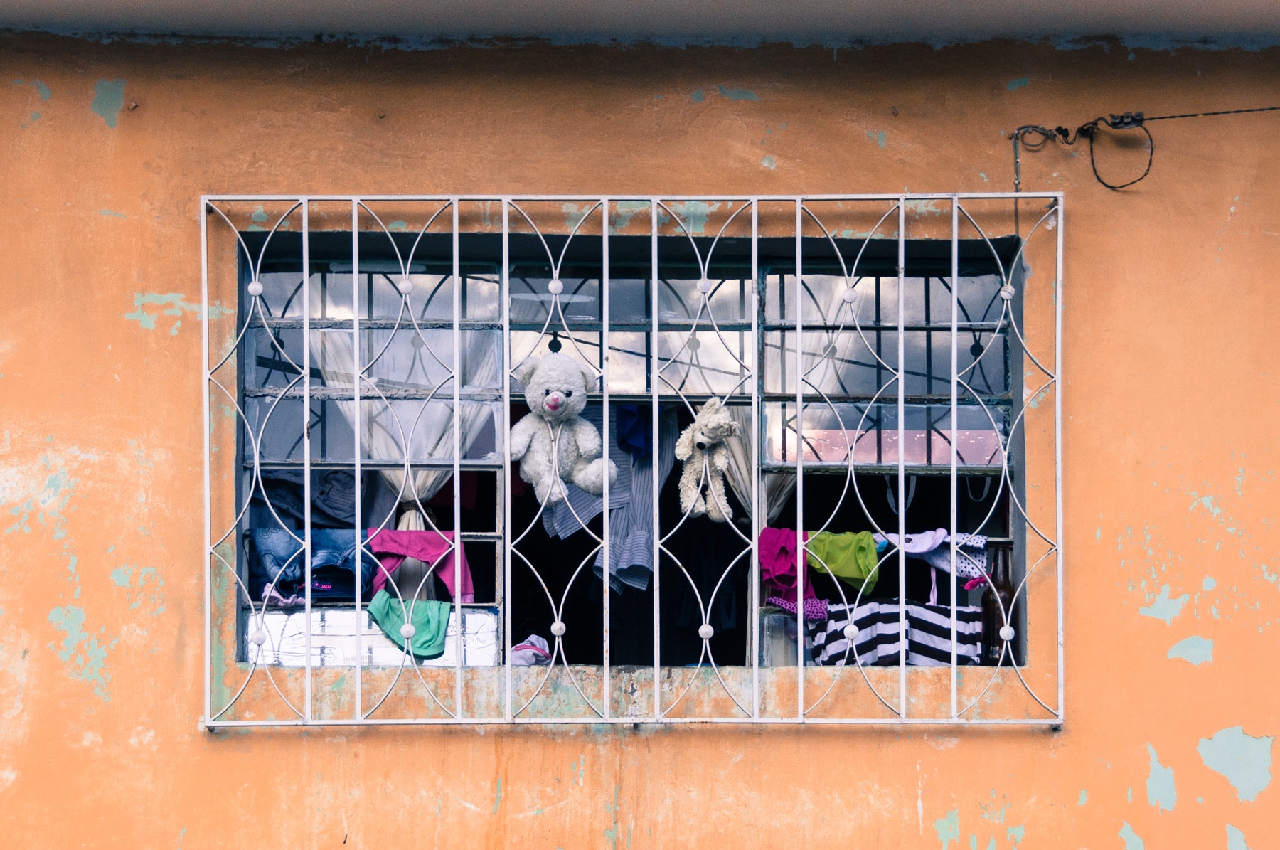 Dos osos de peluche entre ropa y cortinas, detrás de la reja de una ventana en una fachada naranja.