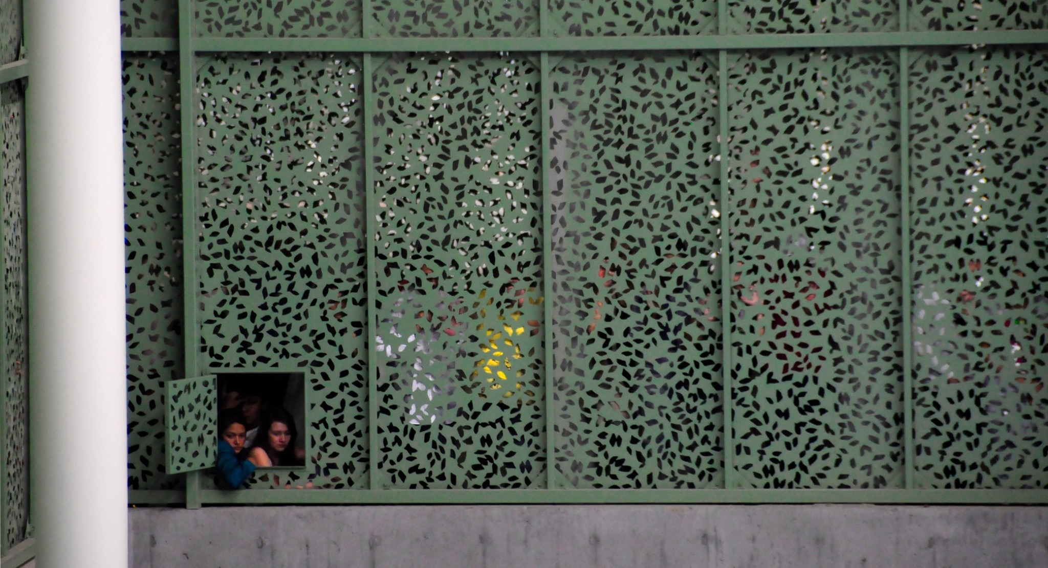 Tres jóvenes observan a través de una pequeña abertura en una fachada verde de patrones perforados.